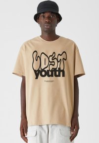 Bežinė medvilninė marškinė su juodu banguotu "LOST youth" piešiniu, trumpomis rankovėmis ir laisvu kirpimu, derinama su šviesiomis kargo kelnėmis ir juoda kibiro kepure.