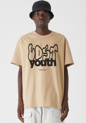 Beige kokvilnas T-krekls ar melnu viļņotu "LOST youth" grafiku, īsām piedurknēm un brīvu piegriezumu, kombinēts ar gaišu krāsu kargo biksēm un melnu bucket cepuri.
