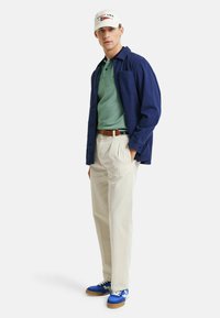 Camisa de botones azul marino, polo verde, pantalones crema y zapatillas deportivas azules. Gorra blanca con detalles en texto rojo y azul. Atuendo casual con texturas variadas.