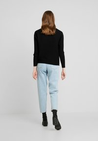 Černý žebrovaný svetr s dlouhými rukávy, kombinovaný se světle modrými zúženými džíny a černými kotníkovými botami. Neutrální pozadí, důraz na detaily oblečení.