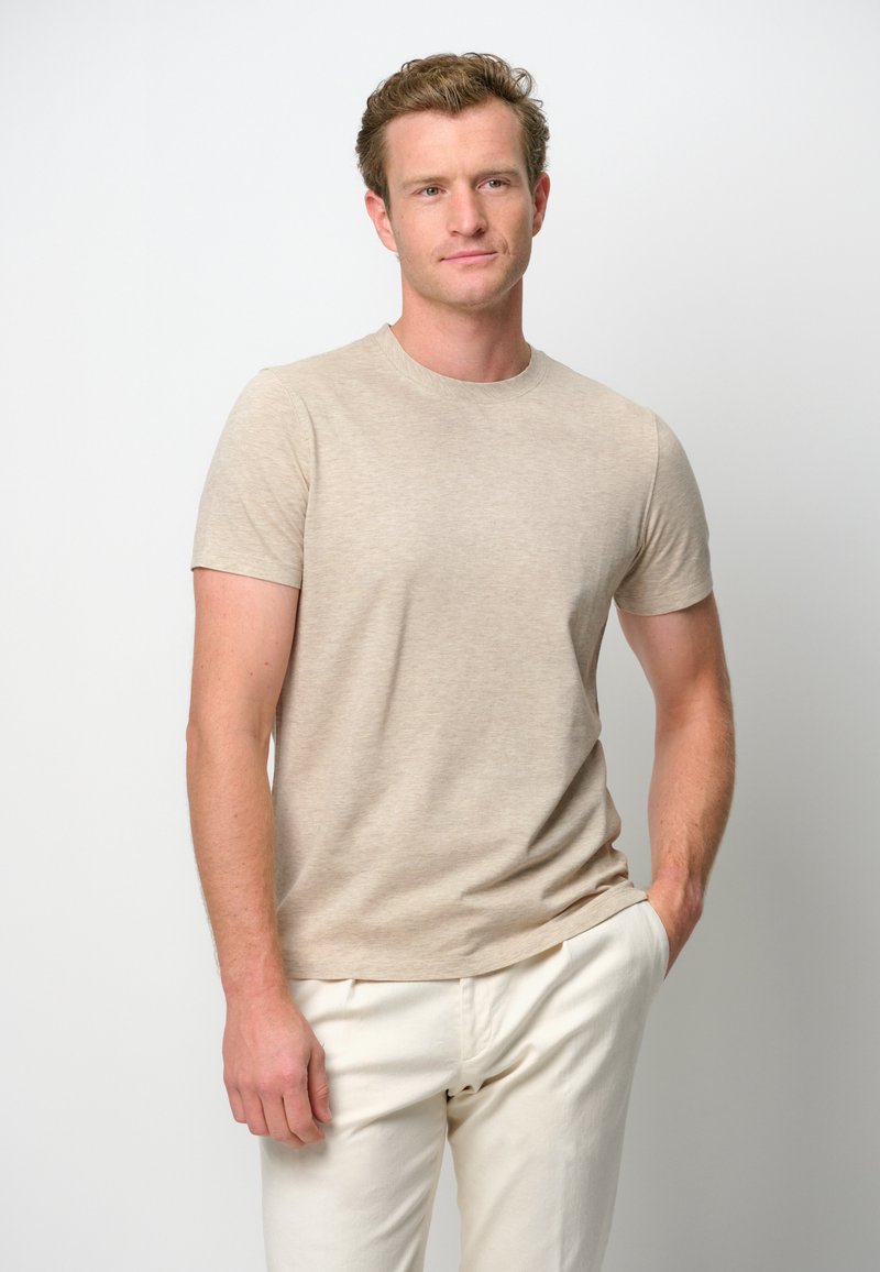 T-shirt beige a maniche corte realizzato in cotone misto, con collo rotondo e texture liscia. Indossato con pantaloni chiari.
