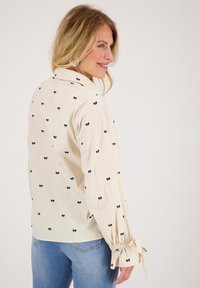 Crème blouse met kleine zwarte strikprints, gerafelde manchetten en een kraag, gecombineerd met lichte blauwe jeans.