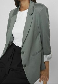 Kobieta w szarym żakiecie, białej koszuli i czarnych spodniach z wiązanym w pasie, siedząca z jedną ręką opartą na drewnianej powierzchni.