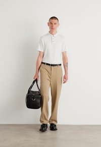 Witte polo met drie knopen, beige broek, zwarte riem, zwarte schoenen en een zwarte tas met een zilveren accent. Staande tegen een effen achtergrond.