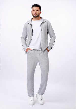 Grauer Sportjackett mit schwarzen Streifenakzenten, kombiniert mit passenden grauen Hosen. Über einem weißen T-Shirt getragen. Weiße Sneaker runden den Look ab.