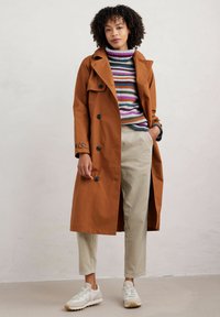 Brązowy płaszcz trench z czarnymi guzikami, noszony na kolorowym swetrze w paski, w połączeniu z beżowymi spodniami z kordurowej tkaniny i białymi trampkami.