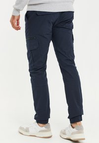 Pantalon cargo bleu marine à bas élastiques, poches latérales et détails zippés. Associé à des baskets blanches et grises avec une semelle épaisse.