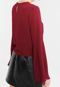 Bordeaux langærmet top med høj hals og åbning i ryggen, parret med en sort skinnende lædernederdel.