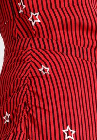 Roter Stoff mit vertikalen schwarzen Streifen und weißen bestickten Sternen. Die Oberfläche wirkt glatt mit seitlich gerafften Details.