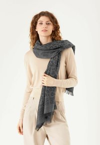 Écharpe gris charbon avec motif léopard, bords frangés. Tissu doux et texturé drape sur un pull beige clair. Style décontracté.