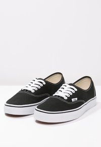 Paire de baskets Vans en toile noire avec lacets blancs et semelles en caoutchouc blanches sur une surface blanche et un fond clair.
