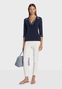 Pull-over en maille bleu marine avec bordure blanche, manches trois-quarts, associé à un pantalon blanc ajusté et des sandales argentées. Porte un sac à main bleu clair.