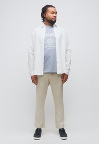 Άνδρας στέκεται φορώντας ένα λευκό ανοιχτό πουκάμισο πάνω από ένα ανοιχτό μπλε t-shirt με γράφημα, μπεζ παντελόνι και μαύρα αθλητικά παπούτσια, κοιτάζοντας προς τα πλάγια.