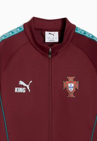 Veste zippée Puma couleur bordeaux avec des rayures turquoise à motifs sur les épaules, écusson de football du Portugal à droite, et "KING" avec logo Puma à gauche.