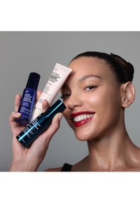 Femme souriante avec un chignon élégant tenant trois produits de soin de la peau près de son visage sur un fond gris uni.