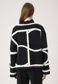 Mujer con cabello largo y marrón viste un suéter negro de felpa con líneas geométricas blancas audaces, mirando hacia atrás contra un fondo claro y liso.