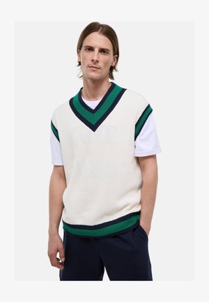 Jeune homme portant un pull sans manches en tricot blanc avec des rayures vertes et bleu marine sur une chemise blanche à manches courtes, debout avec une main dans la poche.