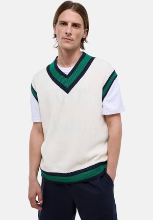 Jeune homme portant un pull sans manches en tricot blanc avec des rayures vertes et bleu marine sur une chemise blanche à manches courtes, debout avec une main dans la poche.