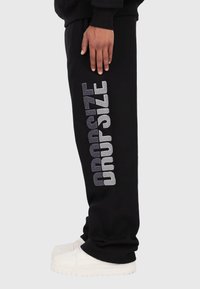 Schwarze Jogginghosen aus weichem Stoff, mit großen, metallisch silbernen "DROPSIZE"-Buchstaben an der Seite. Ausgestattet mit einem elastischen Bund.