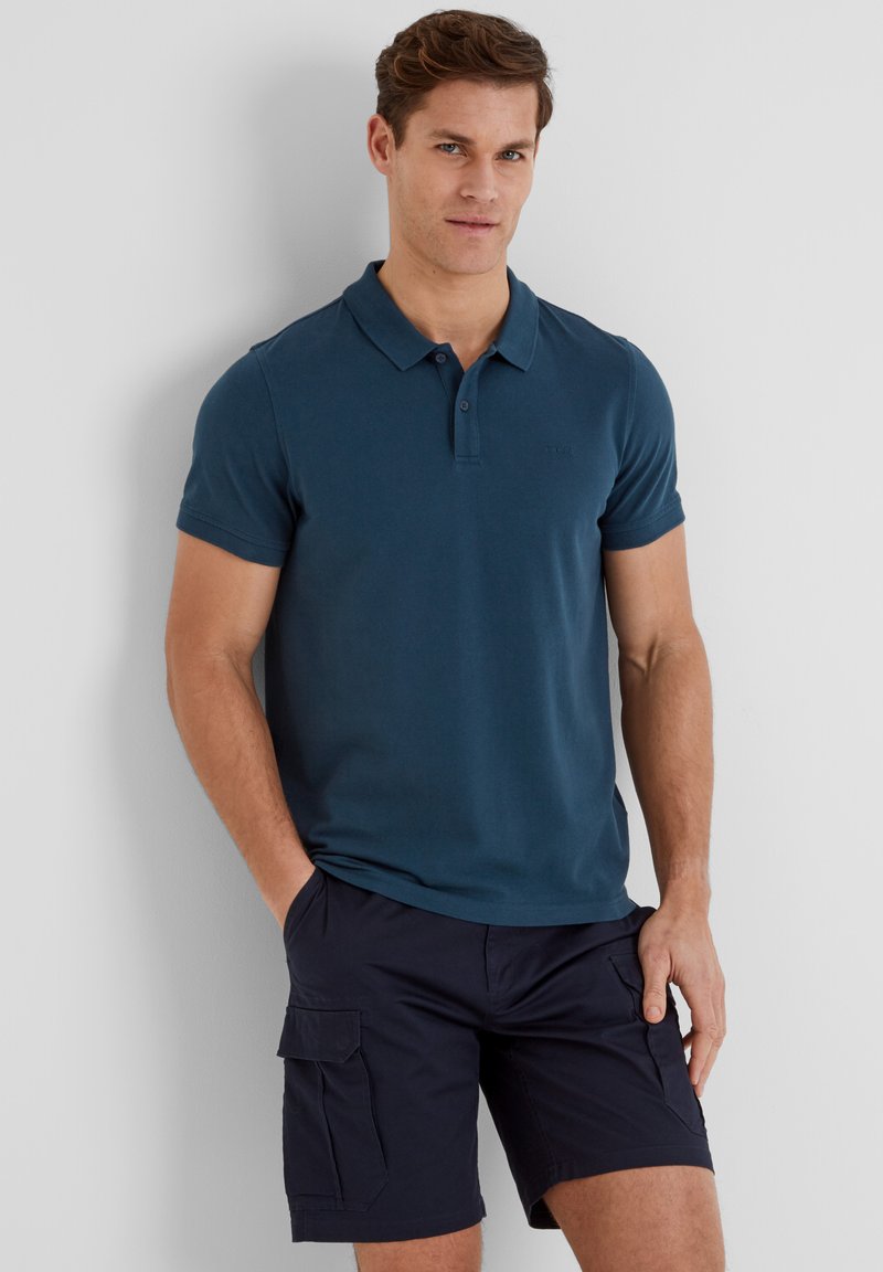 Man standing with hands in pockets wearing a dark blue polo shirt and navy cargo shorts against a plain light background.