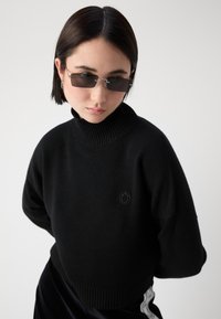 Zwart cropped sweater met een hoge geribbelde kraag en een klein reliëf smiley-logo. Model draagt een donker zonnebril, gestyled met zwarte broeken.
