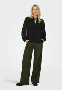 Schwarze, durchsichtige Bluse mit gebundenem Kragen und vertikalen Falten, kombiniert mit weiten, olivgrünen Cordhosen. Schwarze Slipper.