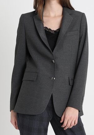Kvinde iført mørkegrå skræddersyet blazer over sort blonde top og mørke ternede bukser, stående mod lysegrå baggrund.