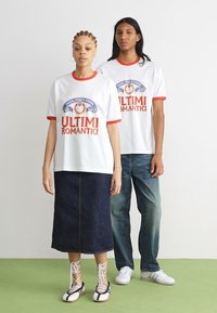 Witte T-shirts met rode accenten en grafische prints, gecombineerd met een lange spijkerrok en blauwe jeans. Schoenen zijn wit en zwart met veters.
