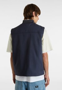 Gilet senza maniche blu navy realizzato in un tessuto resistente, caratterizzato da un colletto alto e bordi puliti, abbinato a una camicia a righe di colore chiaro sottostante.