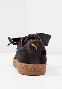 Černé tenisky Puma s gumovou podrážkou viděné ze zadu, s zlatým logem Puma na patní části a širokými černými saténovými tkaničkami.