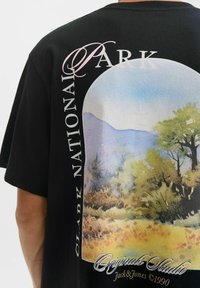 Melna T-krekla aizmugure ar krāsainu dabas skatu, kas ietver kokus, kalnus un tekstu "Klārka Nacionālais parks" un "Originals Studio Jack & Jones."