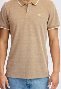Tan polo shirt made of textured material, featuring a ribbed collar with cream accents, two buttons, and a small logo on the chest.