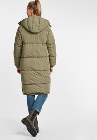 Olivgrön puffjacka med horisontella sömmar, huva och längre längd, matchad med blå jeans och bruna ankelstövlar.