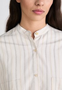Camicia bianca con bottoni, caratterizzata da righe verticali in nero e marrone chiaro, con colletto alla cinese e tasca piatta sul lato sinistro.