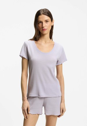Femme portant un haut violet clair à manches courtes et un short assorti en tissu texturé, debout devant un fond blanc uni.
