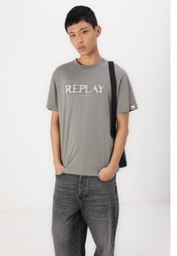 Grey short-sleeve t-shirt featuring "REPLAY" logo in a slightly distressed font. Paired with dark grey denim jeans and a black bag strap.