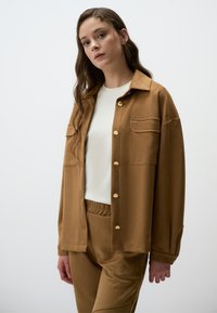Veste chemise en tissu doux marron avec un col, boutons dorés et une poche poitrine, portée avec un pantalon marron assorti et une chemise blanche.