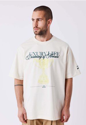 Man with short hair and beard wearing white oversized T-shirt with "LUXURY LIFE Dreaming of Monaco" and a yellow trophy graphic.