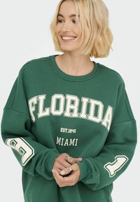 Felpa verde con "FLORIDA" in lettere bianche in grassetto, con "MIAMI" e l'anno di fondazione. Maniche larghe, tessuto morbido.