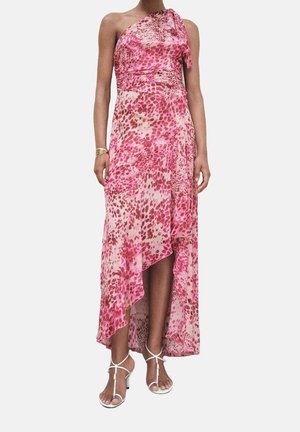 Kobieta w różowo-czerwonej asymetrycznej sukience na jedno ramię z abstrakcyjnym wzorem oraz białych sandałach na obcasie na gładkim tle.