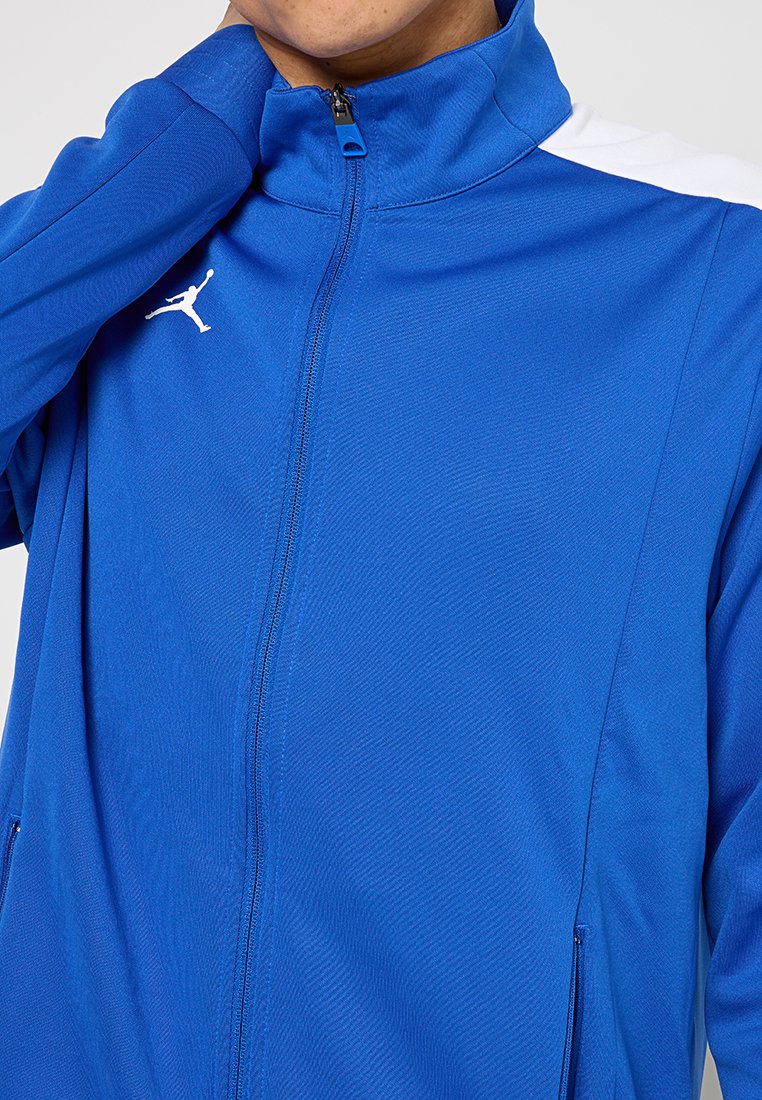Person wearing a blue zip-up sports jacket with white logo on chest and a white stripe on the shoulder, hand touching neck.