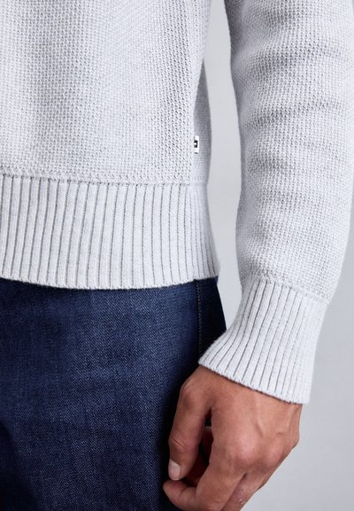 Lichtgrijze gebreide trui met ribbelmanchetten en -zoom, met een gestructureerd weefpatroon. Hand zichtbaar die de trui tegen donkere denim houdt.