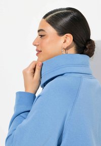 Felpa blu con colletto alto, polsini a coste e una texture morbida. Presenta una vista laterale e una mano che tiene il colletto.