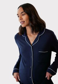 Granatowa koszula od piżamy z białymi lamówkami, z kołnierzykiem, zapinana na guziki, z kieszonką na piersi i długimi rękawami, uszyta z miękkiego materiału.