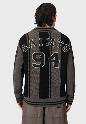 Mężczyzna ubrany w czarno-szary sweter w paski z napisem "SAINTS 94" na plecach, w połączeniu z luźnymi szarymi spodniami.