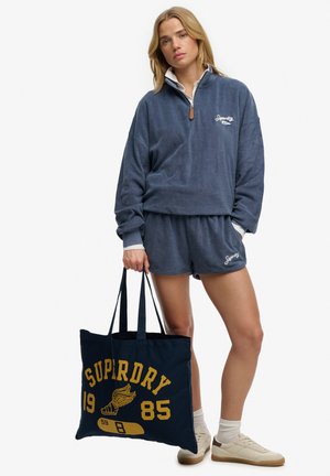 Donna che indossa un pullover in pile blu navy Superdry e pantaloncini, tiene una borsa tote blu navy con il logo Superdry giallo e sneakers bianche.