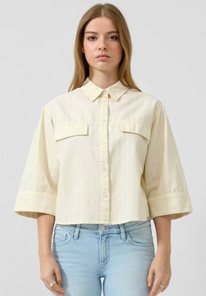 Jeune femme portant une chemise courte boutonnée jaune pâle à manches larges et un jean bleu clair, sur un fond uni.