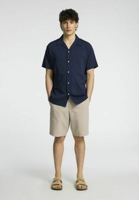 Camisa de botão de manga curta azul-marinho com colarinho, combinada com calções bege. O modelo está em pé com as mãos nos bolsos, usando sandálias castanho-claro.