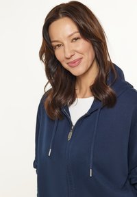 Felpa zip con cappuccio di colore blu navy, dotata di coulisse, tasche laterali e una texture liscia. Presenta una zip argentata e un design con vestibilità comoda.