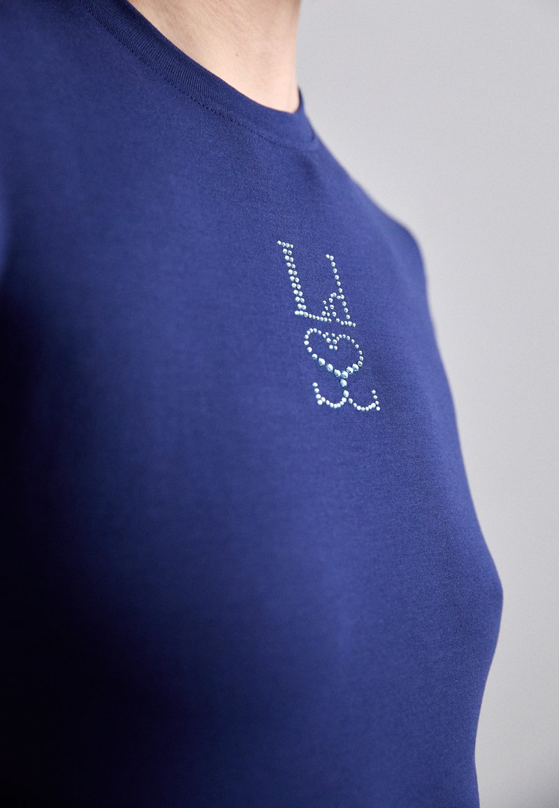 Navy blue cotton t-shirt with a fitted design, featuring a rhinestone-embellished logo on the front for decorative detail.